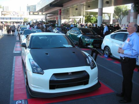 1998 Ford Crown Victoria - white Nissan GT-R with a black carbon-fiber hood leads a row of show cars on red flooring as a man stands beside it.