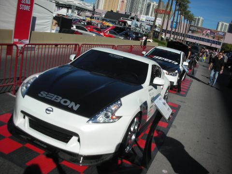 1998 Ford Crown Victoria - white Nissan 370Z fitted with a black SEIBON carbon hood sits on a checkered display floor at an outdoor show.