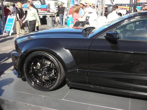 1998 Ford Crown Victoria - Side profile of a dark muscle coupe with black multi-spoke wheels on a display platform at a show.