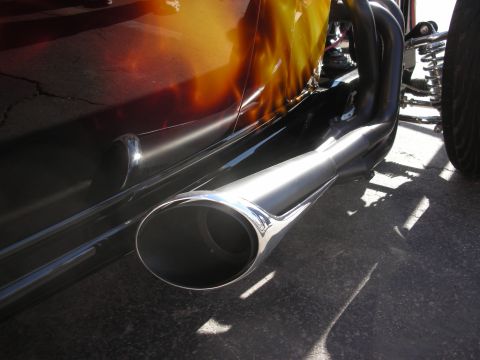 1998 Ford Crown Victoria - Low-angle close-up of a polished chrome exhaust tip beneath a hot rod's fire-flame painted body.