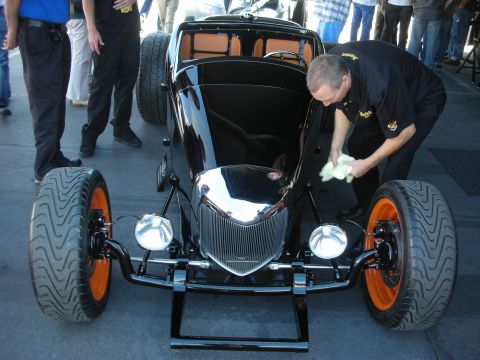 1998 Ford Crown Victoria - man polishes the chrome grille and black bodywork of a fenderless hot rod displayed on a stand at an indoor event.