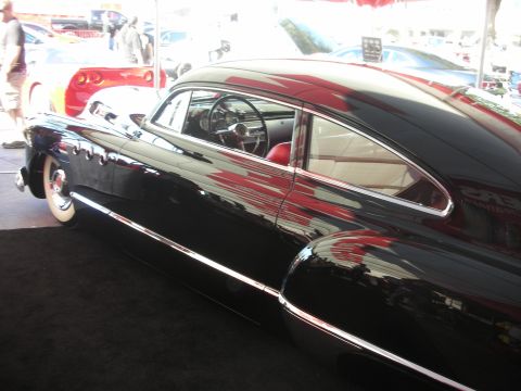 1998 Ford Crown Victoria - Side view of a black chopped fastback custom sedan with whitewall tires and red interior, displayed under a tent.