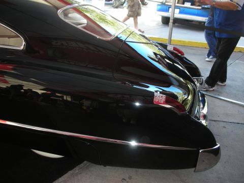 1998 Ford Crown Victoria - Rear quarter detail of a glossy black custom car showing a chrome bumper, body emblem and a person walking past.