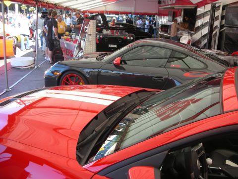 1998 Ford Crown Victoria - red sports car with white stripes in the foreground and a black coupe with red wheels at a busy outdoor car show.