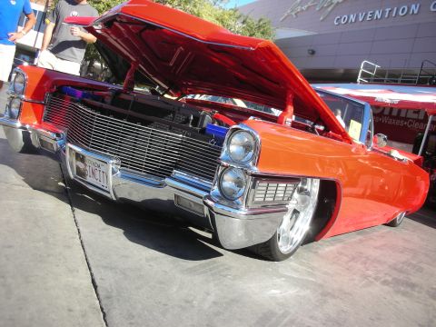 1998 Ford Crown Victoria - orange lowered custom Cadillac with chrome bullet bumpers and a billet grille sits hood-up near a convention center.