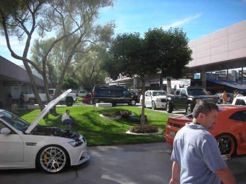 1998 Ford Crown Victoria - landscaped expo courtyard with a white hooded-open car, an orange coupe and parked trucks as visitors mill about.