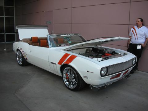 1998 Ford Crown Victoria - white first-generation-style Camaro convertible with orange stripes and open hood on display beside a building.