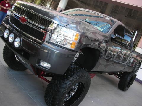 1998 Ford Crown Victoria - lifted gray Chevrolet pickup truck with ghost-flame airbrush paint, light bar and large mud-terrain tires.