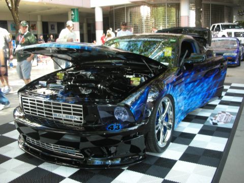 1998 Ford Crown Victoria - black Saleen Mustang with blue flame graphics and chrome wheels sits on a checkered display floor at a show.