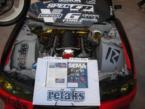 1998 Ford Crown Victoria - Engine bay of a red car showing a swapped V8 with sponsor banner and a SEMA info sheet propped on the body.