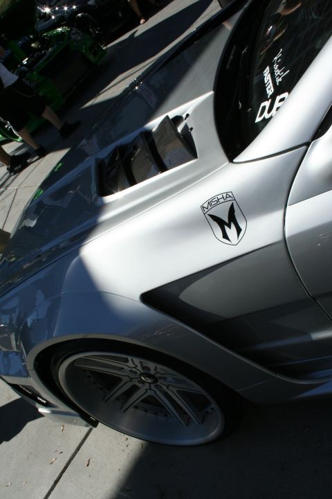 1998 Ford Crown Victoria - Side detail of a silver sports car showing a Misha M-logo decal near the front fender vent above a dark multi-spoke wheel.