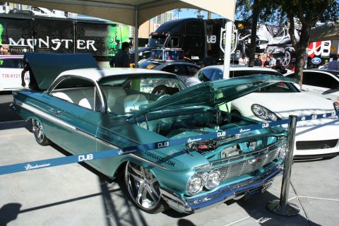 1998 Ford Crown Victoria - teal custom Chevrolet Impala with hood up sits on polished wheels among Budweiser and DUB barriers at an outdoor show.