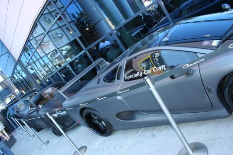 1998 Ford Crown Victoria - Profile view of a matte gray car wearing Giovanna and Specialty Car Craft decals next to a glass-walled building.