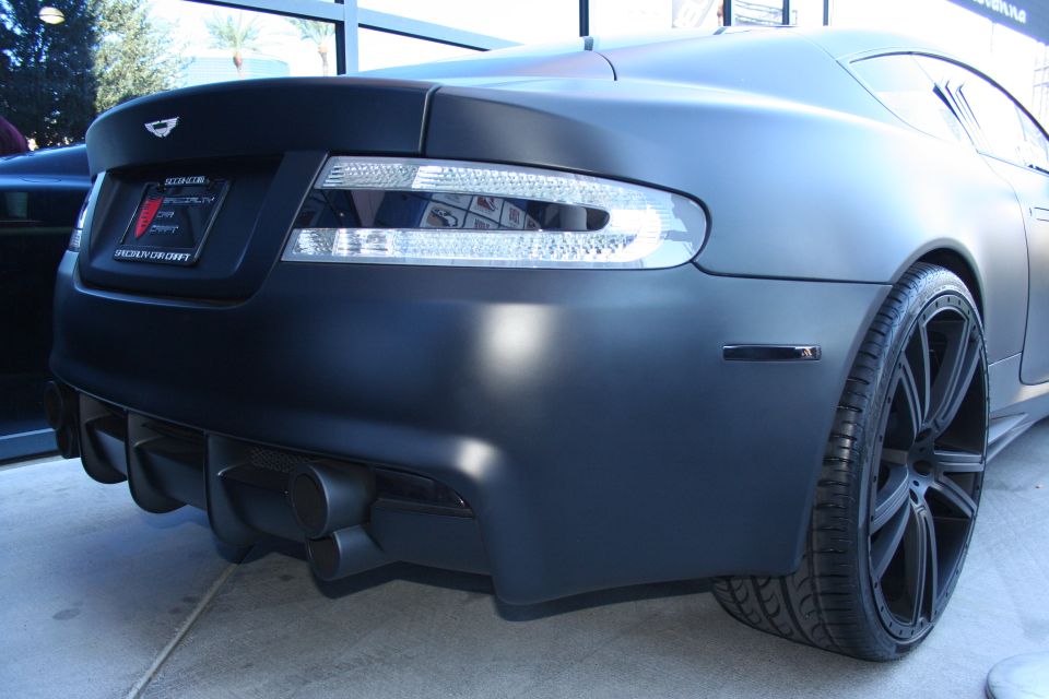 Rear of a matte black Aston Martin coupe showing tail lights, diffuser and black wheel