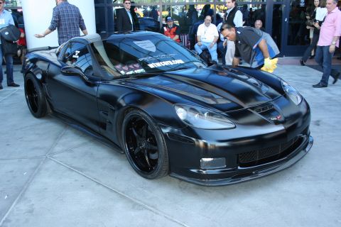 1998 Ford Crown Victoria - Black Chevrolet Corvette C6 with a widebody kit and black wheels surrounded by people at an outdoor show.