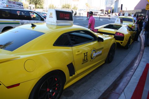 1998 Ford Crown Victoria - Yellow Chevrolet Camaro with Top Gear and History decals and roof sign parked beside a yellow Lamborghini at SEMA.