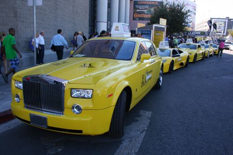 1998 Ford Crown Victoria - Yellow Rolls-Royce Phantom sedan with a roof sign leading a row of yellow promotional cars along a busy street.