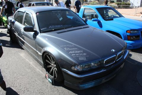 1998 Ford Crown Victoria - lowered gray BMW 7 Series sedan covered in sponsor decals sits on dark wheels at an outdoor automotive show.