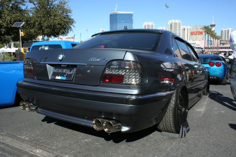 1998 Ford Crown Victoria - Rear three-quarter view of a gray BMW 7 Series sedan with dual chrome exhaust tips, parked before a city skyline.