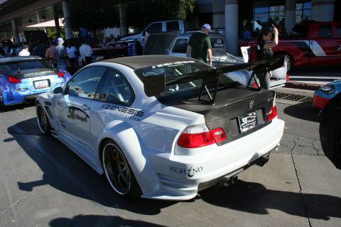 1998 Ford Crown Victoria - white widebody BMW M3 coupe with a tall carbon-fiber rear wing and sponsor graphics, seen from the rear quarter.