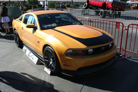 1998 Ford Crown Victoria - orange Ford Mustang coupe with a black hood stripe and dark wheels displayed on pavement behind a barrier.