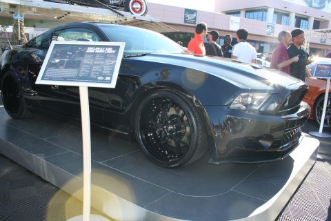 1998 Ford Crown Victoria - glossy black Ford Mustang on black wheels sits on a display platform with an info placard at an outdoor event.