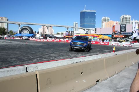 1998 Ford Crown Victoria - blue Ford F-150 Raptor sits on a dirt-and-asphalt demo course ringed by Lucas Oil banners, with Las Vegas hotel towers in the background.