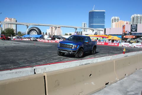 1998 Ford Crown Victoria - blue Ford F-150 Raptor moves across a paved demo course lined with Lucas Oil and SEMA banners under a clear blue sky.