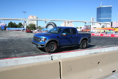1998 Ford Crown Victoria - Front three-quarter view of a blue Ford F-150 Raptor on a dirt demo course with event banners and Vegas buildings behind.