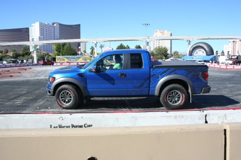 1998 Ford Crown Victoria - Side profile of a blue Ford F-150 Raptor with graphic decals, parked on an asphalt course with a concrete barrier in front.