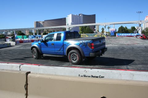 1998 Ford Crown Victoria - Rear three-quarter view of a blue Ford F-150 Raptor with side graphics on a paved course, Vegas hotels behind.