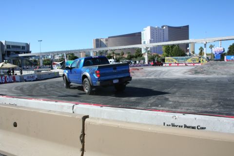 1998 Ford Crown Victoria - blue Ford F-150 Raptor with bold side graphics drives away across a paved demo course toward distant Las Vegas hotels.