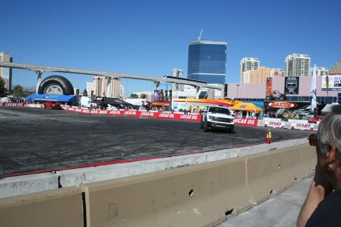 1998 Ford Crown Victoria - white pickup truck sits at the far edge of a paved demo course lined with Lucas Oil banners and a giant tire display, Vegas towers behind.