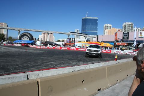 1998 Ford Crown Victoria - white pickup truck on a wide asphalt demo course surrounded by Lucas Oil and SEMA banners under a clear sky.