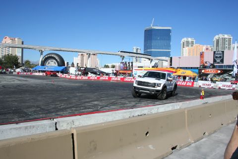 1998 Ford Crown Victoria - white Ford F-150 Raptor seen front three-quarter on a paved course ringed by Lucas Oil banners, Vegas buildings behind.