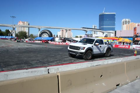 1998 Ford Crown Victoria - white Ford F-150 Raptor with dark side graphics on a paved demo course, monorail bridge and Vegas hotels in the background.