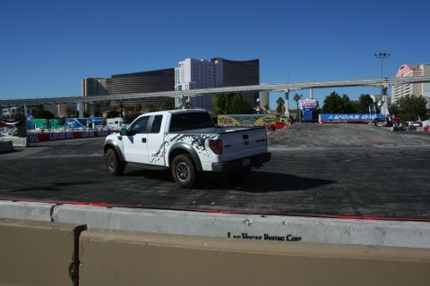 1998 Ford Crown Victoria - Rear three-quarter view of a white Ford F-150 Raptor with side graphics on a paved course, Vegas hotels behind.
