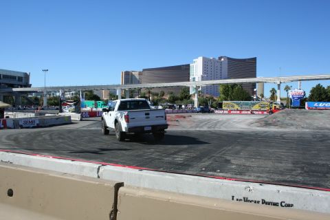 1998 Ford Crown Victoria - white pickup truck drives across a broad asphalt demo course ringed by banners, with Vegas towers and a monorail bridge behind.