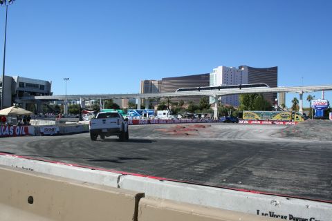 1998 Ford Crown Victoria - white pickup truck on a wide asphalt demo course ringed by event banners, with a monorail bridge and Vegas towers behind.