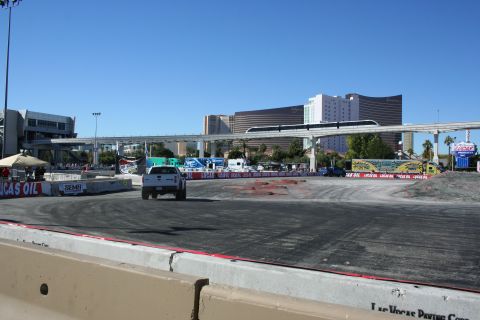 1998 Ford Crown Victoria - small white pickup truck sits on a broad asphalt demo course lined with banners, Las Vegas hotels in the background.
