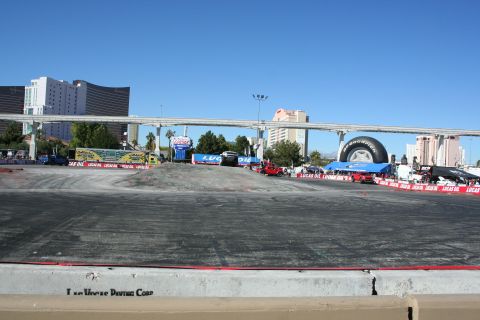 1998 Ford Crown Victoria - wide empty paved demo course lined with banners and a giant tire display, monorail bridge and Vegas towers behind.