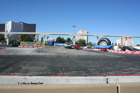 1998 Ford Crown Victoria - paved demo course ringed by Lucas Oil banners with parked trucks and a tire display, monorail bridge and Vegas hotels behind.