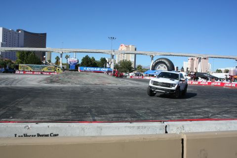 1998 Ford Crown Victoria - white Ford F-150 Raptor drives along the edge of a wide paved demo course ringed by Lucas Oil banners, Vegas towers behind.