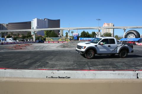1998 Ford Crown Victoria - white Ford Raptor pickup with graphic decals stands on a dirt course bordered by Lucas Oil banners under a clear blue Las Vegas sky.