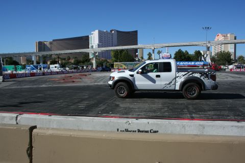 1998 Ford Crown Victoria - white Ford Raptor pickup with black side graphics drives across a dirt course lined with Lucas Oil banners, casino towers behind.