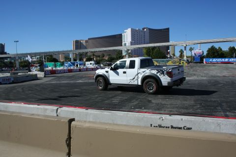 1998 Ford Crown Victoria - Rear three-quarter view of a white Ford Raptor pickup on a graded dirt course with Lucas Oil and SEMA banners and the Las Vegas skyline beh…