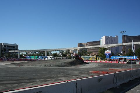 1998 Ford Crown Victoria - empty graded dirt course ringed with Lucas Oil banners under a clear blue sky, with Las Vegas casino towers and a monorail bridge behind.