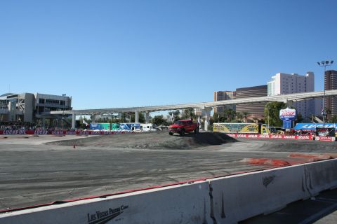 1998 Ford Crown Victoria - small red car crosses a graded dirt course lined with Lucas Oil banners, the Las Vegas skyline and monorail bridge behind.