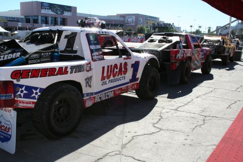1998 Ford Crown Victoria - row of off-road race trucks, the lead one in General Tire and Lucas Oil number-1 livery, lined up on pavement at an outdoor event.