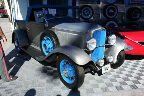 1998 Ford Crown Victoria - bare-metal 1930s-style roadster hot rod with bright blue wire wheels and dual headlights sits on a checkered floor at an indoor show.